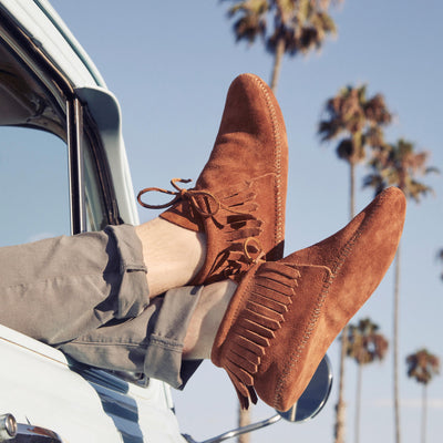 Minnetonka men's classic softsole fringe boot in brown soft suede with rawhide laces and padded insole shown worn outdoors with palm trees background