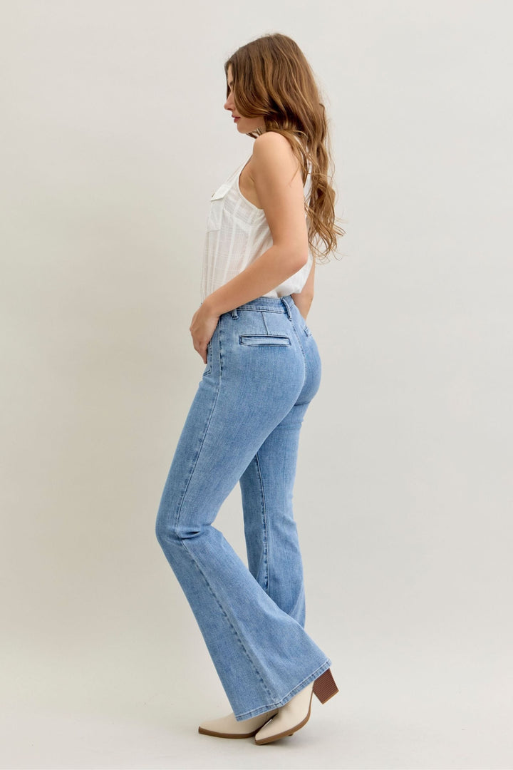 Side view of a woman wearing high-rise flare Judy Blue Jeans in light blue denim with front patch and welt pockets, paired with a white sleeveless top and beige boots.