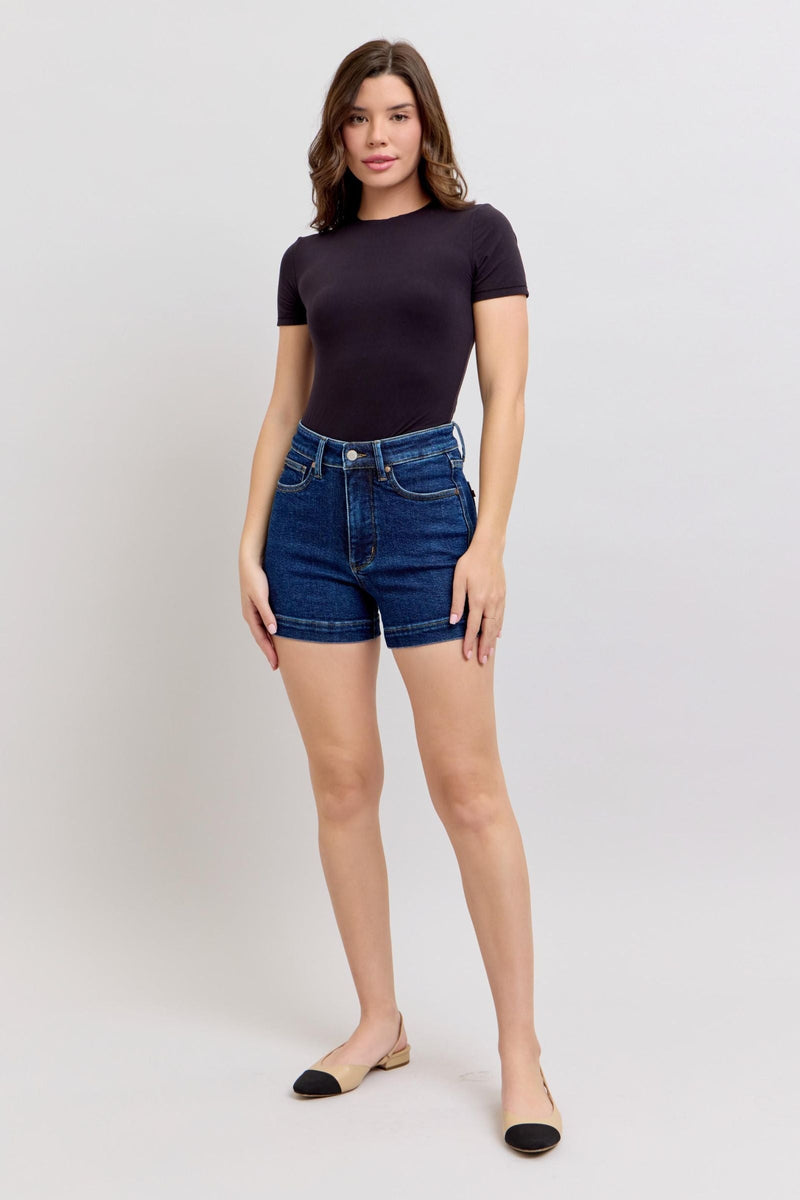 Woman wearing high waist Judy Blue Shorts 150333 in dark blue denim with tummy control and short inseam paired with black fitted top and flats