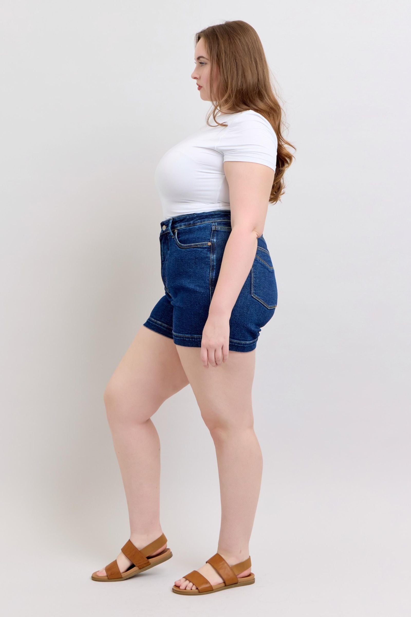 Plus size model wearing high waist Judy Blue Shorts in dark blue denim with tummy control and 4.25 inch inseam paired with a white fitted t-shirt and brown sandals.