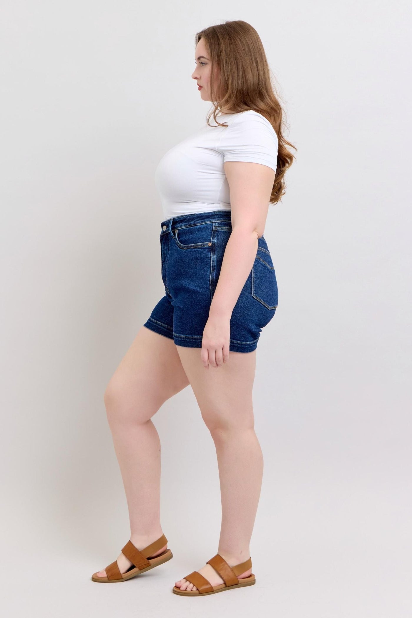 Plus size model wearing high waist Judy Blue Shorts in dark blue denim with tummy control and 4.25 inch inseam paired with a white fitted t-shirt and brown sandals.
