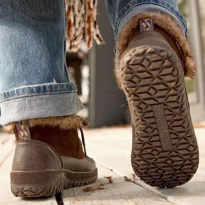 A person wearing Minnetonka Tinley Shoe with suede and PU material, featuring a durable MinnTREAD rubber outsole with rice husk, walking on a wooden surface.