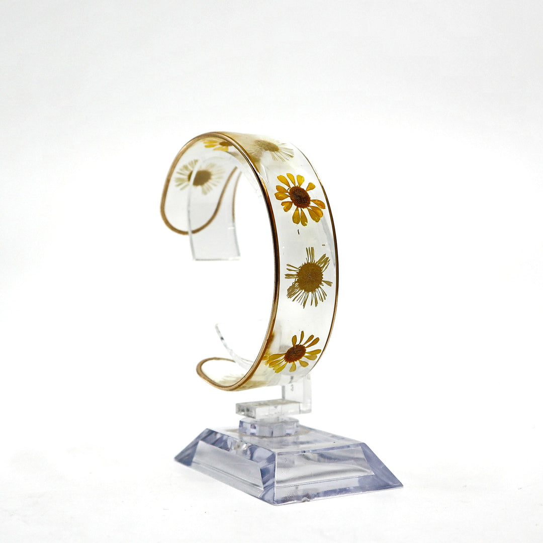 Dried flower resin cuff bracelet with real yellow and white flowers encased in clear resin on a gold-edged band displayed on a clear stand