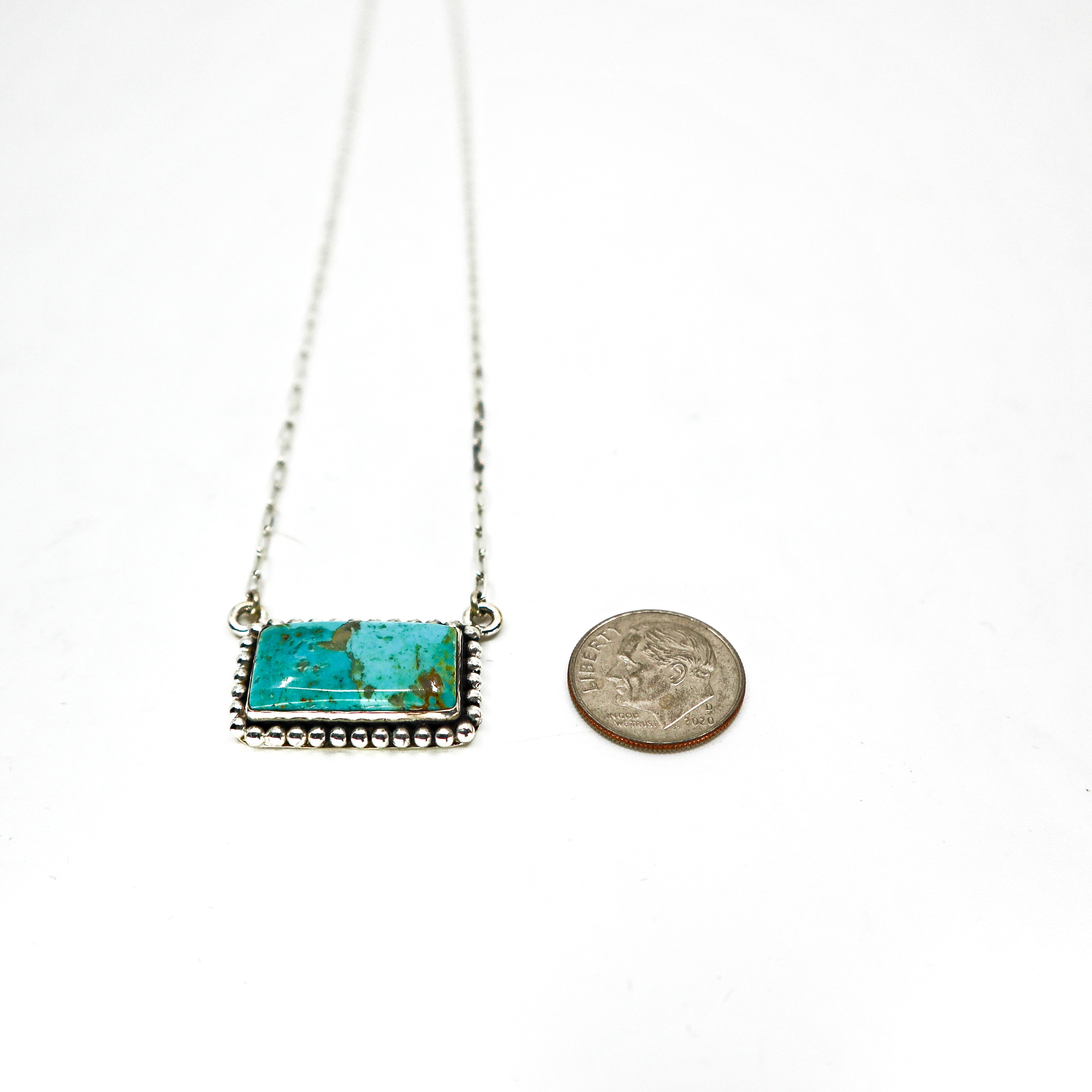 Handmade sterling silver necklace with large turquoise bar pendant and intricate border design next to a dime for size comparison