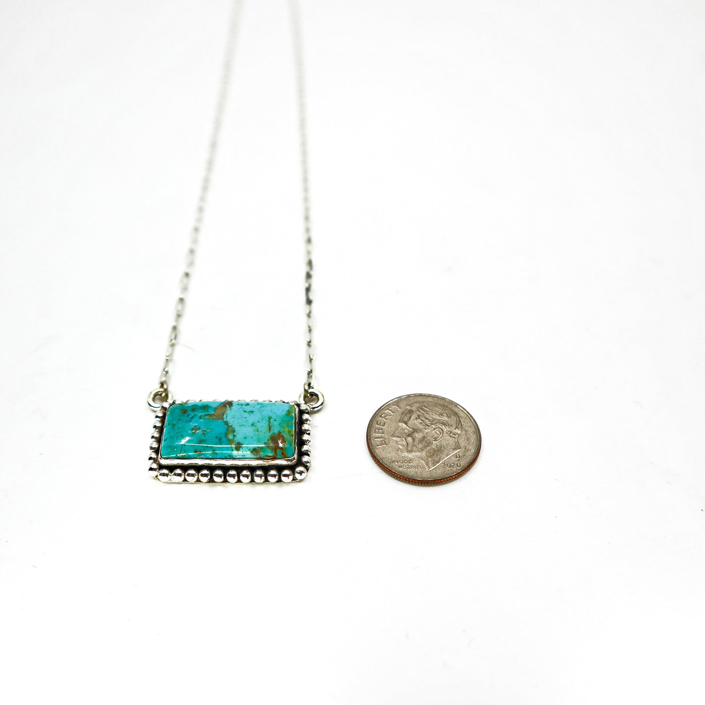 Handmade sterling silver necklace with large turquoise bar pendant and intricate border design next to a dime for size comparison