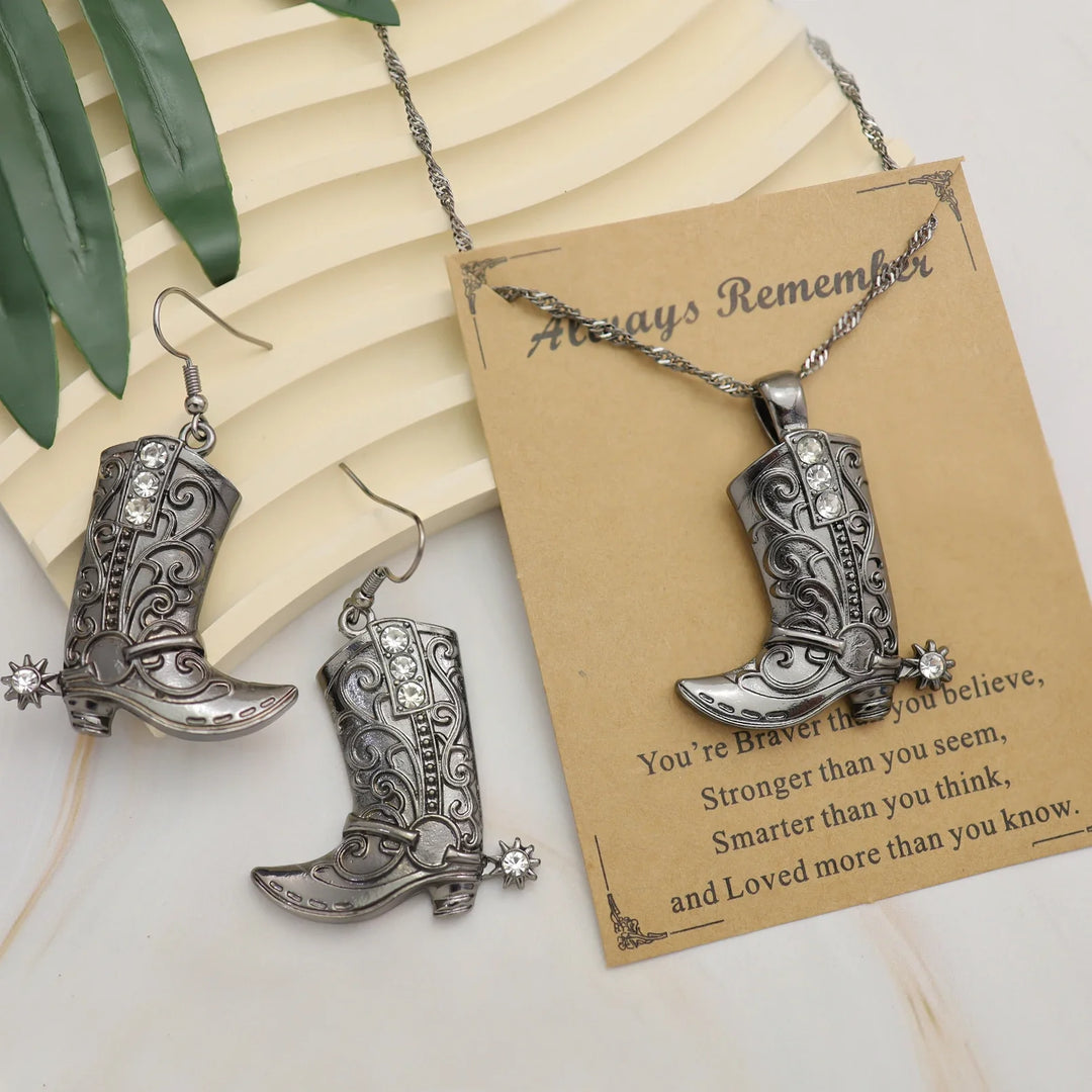 Silver Western cowgirl boot pendant necklace and matching earrings set with intricate detailing and spurs on a rustic card background.